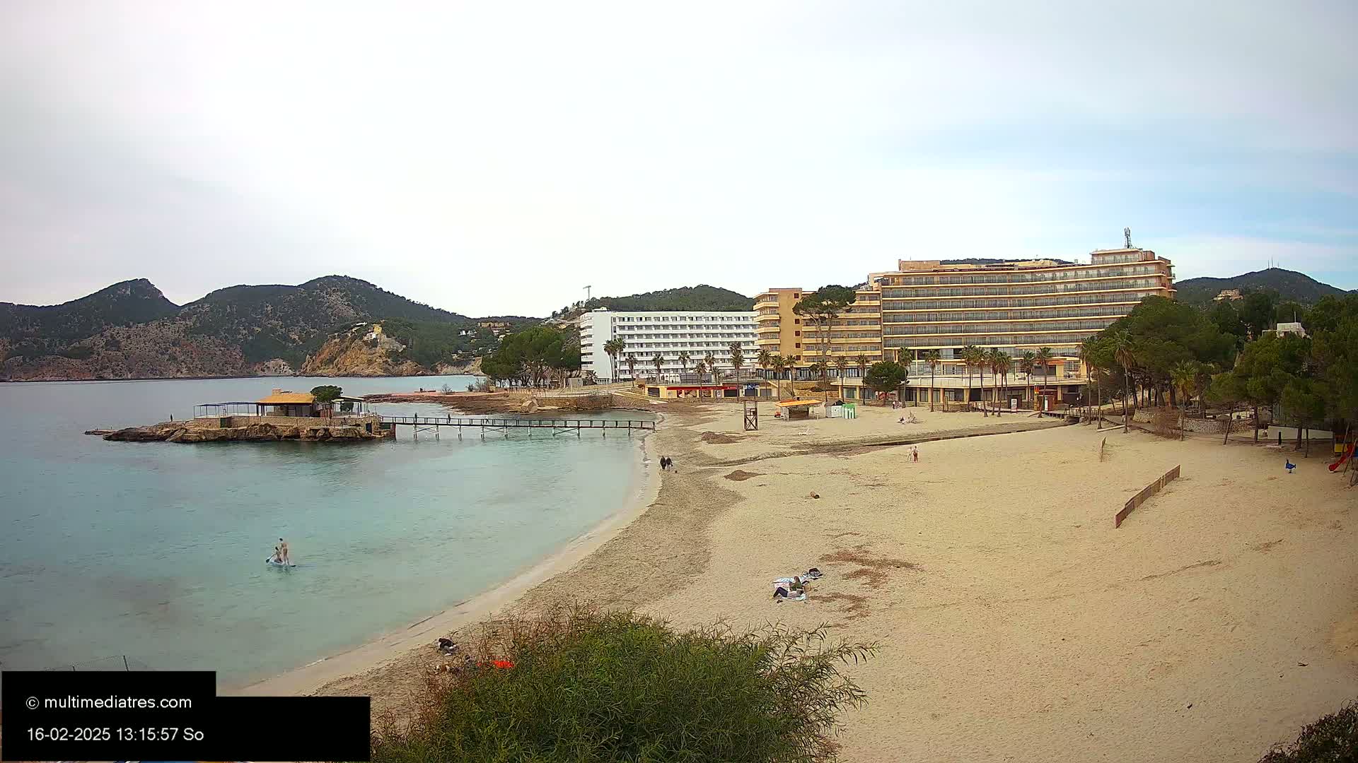 A sandy beach with calm, light-blue water stretches out before two large hotels, backed by green hills under a mostly cloudy sky.