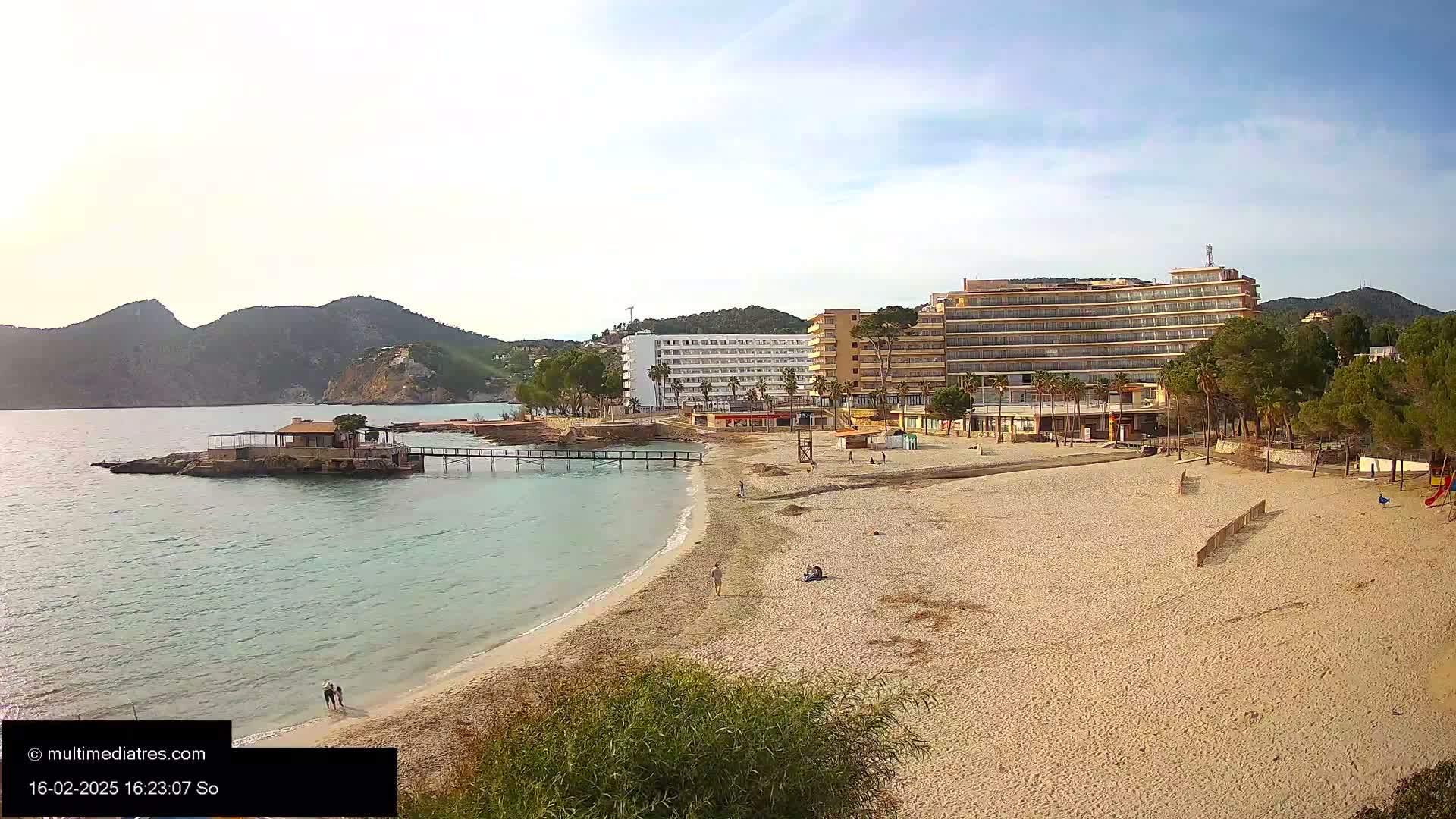 A sandy beach with a few people on it sits next to calm, light-blue water, a small pier, and several large hotels, all backed by green hills under a mostly clear sky.