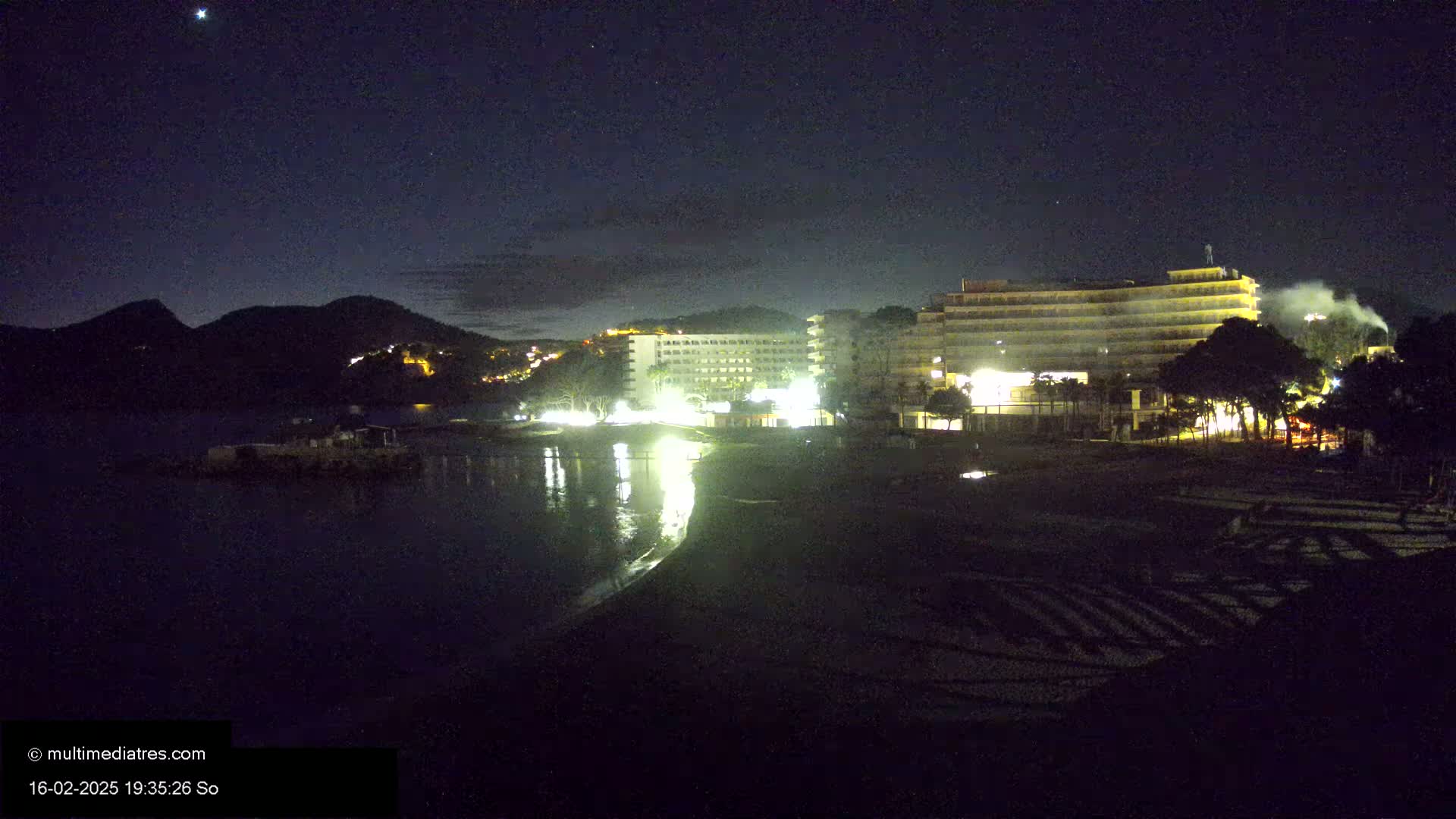 A nighttime view of a beach with brightly lit hotels and a dark, mountainous backdrop under a clear, starry sky.