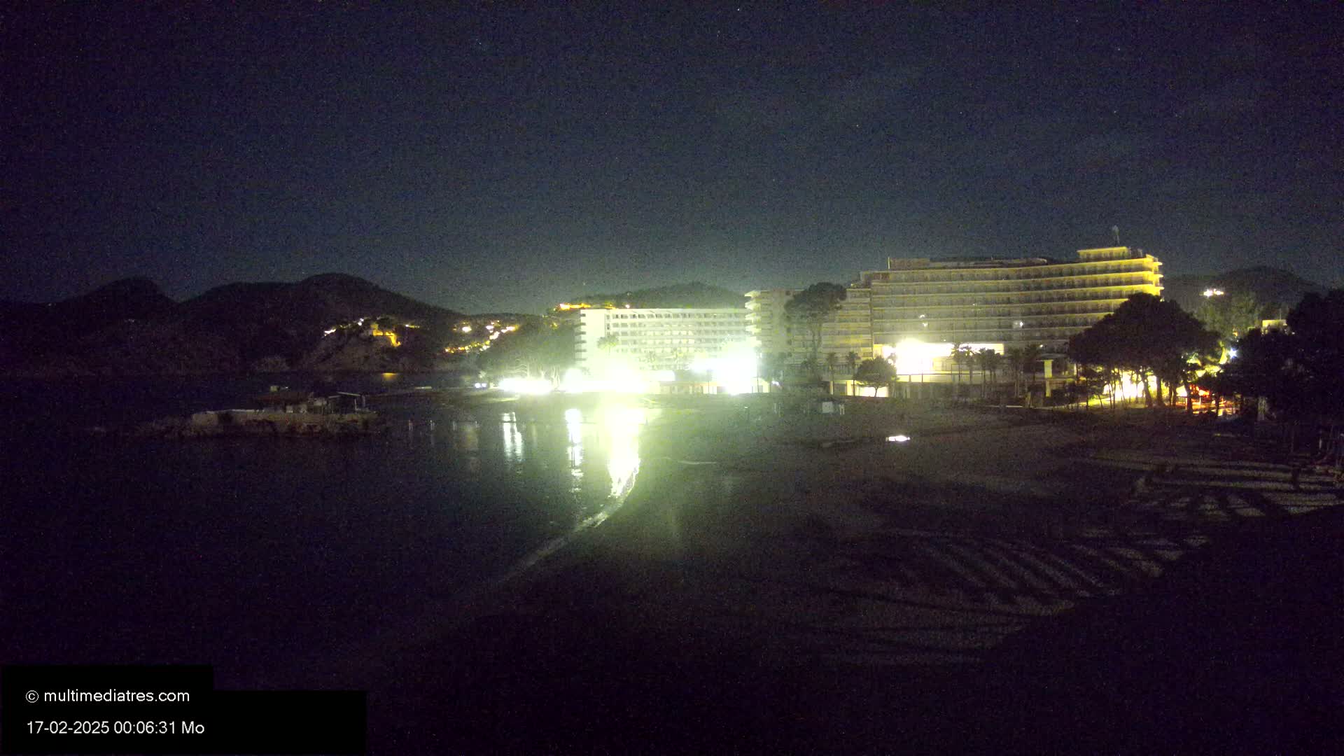 At night, two brightly lit hotels stand on a beach next to a dark body of water and a hill under a clear, starry sky.