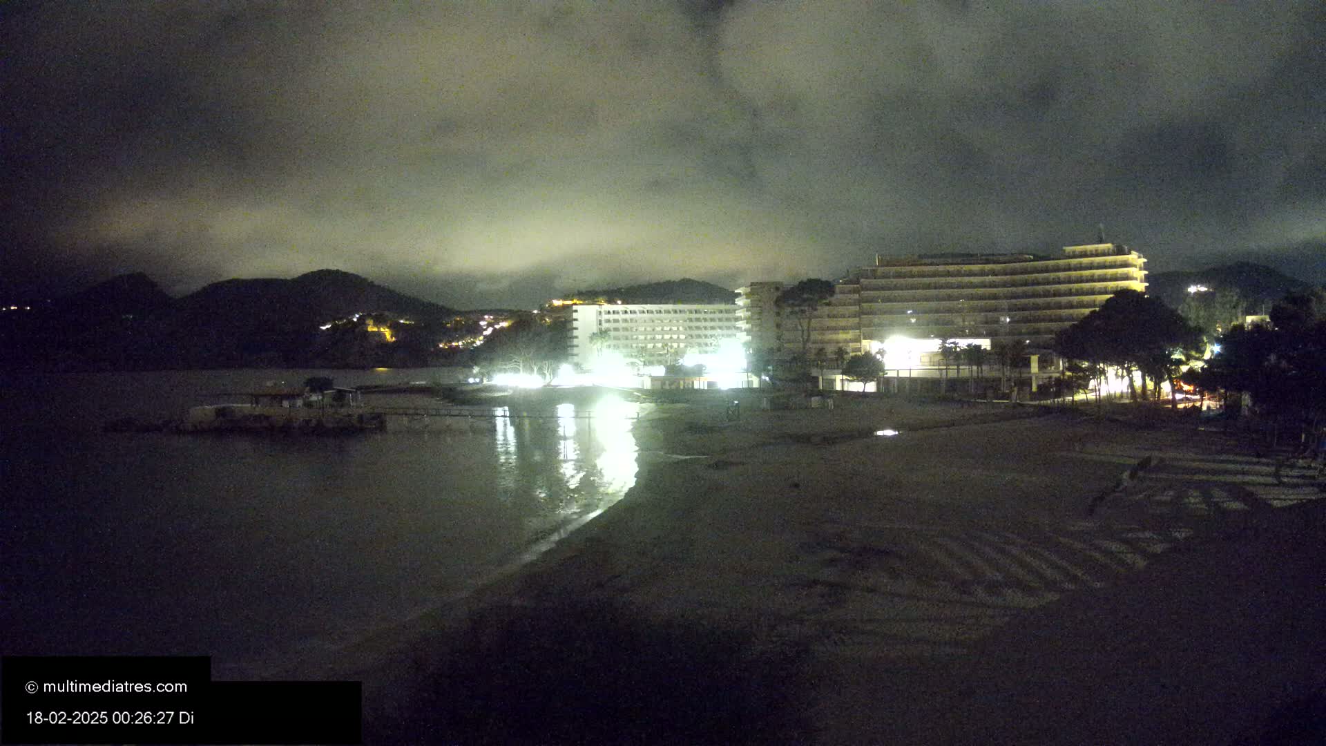 A nighttime view of a beach with two illuminated hotels, a dark mountain in the background, and calm water reflecting the building's lights under a cloudy sky.