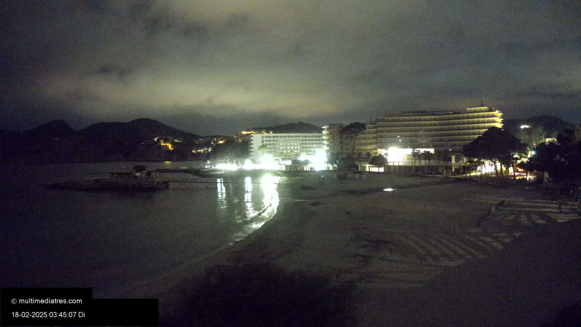 At night, under a cloudy sky, illuminated hotels sit on a beach next to a dark body of water and a hill in the distance.