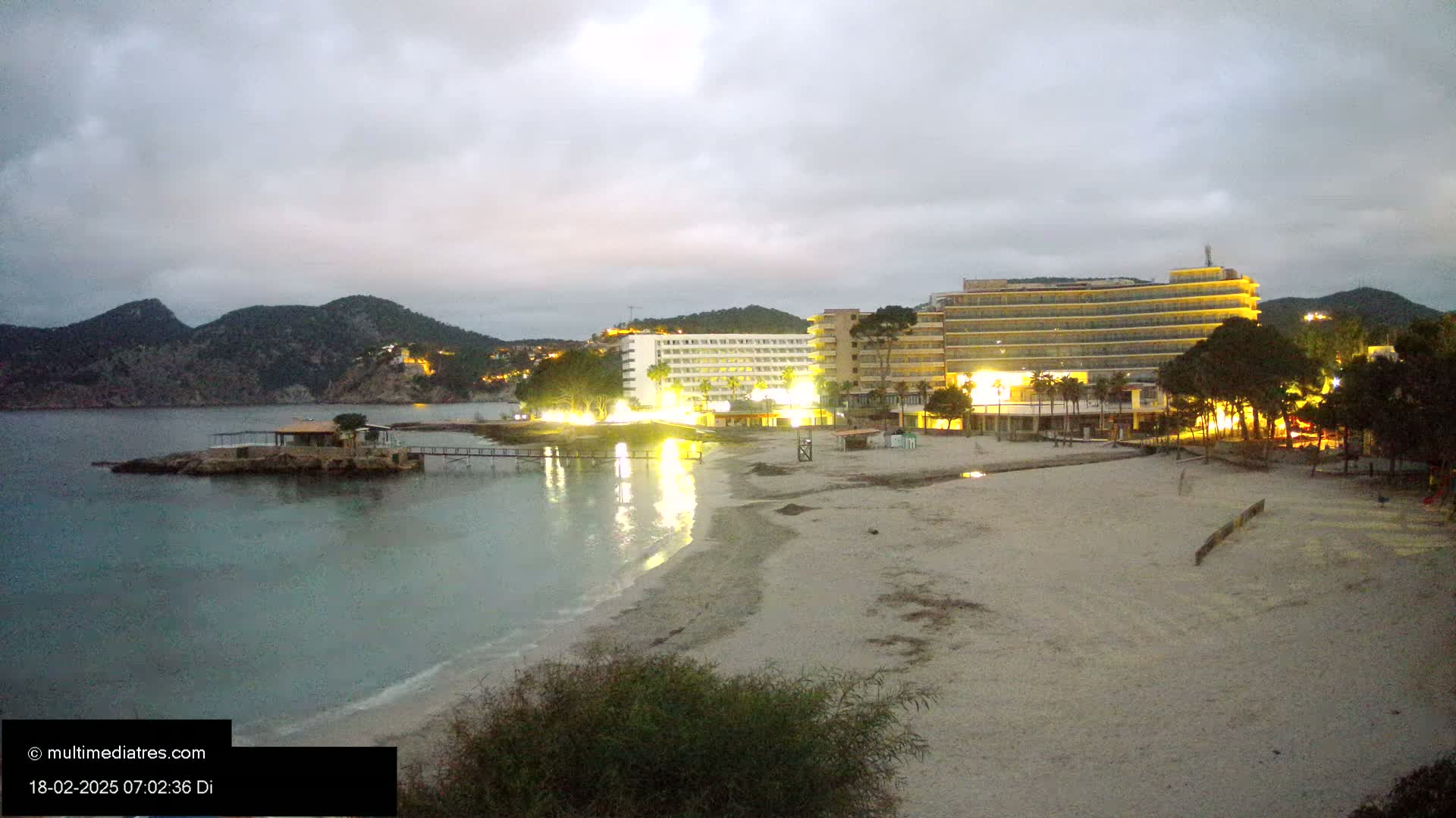 A sandy beach at twilight is illuminated by the lights of hotels and buildings on a calm bay, with dark mountains in the background under a cloudy sky.