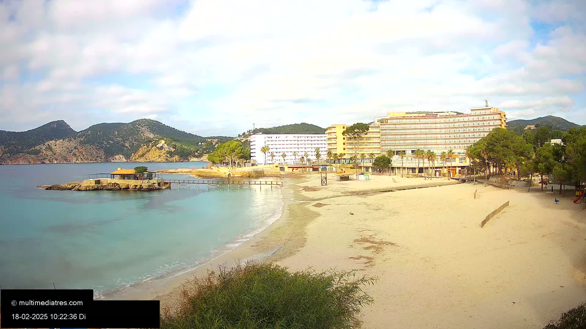 A sandy beach with calm, turquoise water, a small restaurant on a rocky outcropping, and large hotels behind it, all under a partly cloudy sky.
