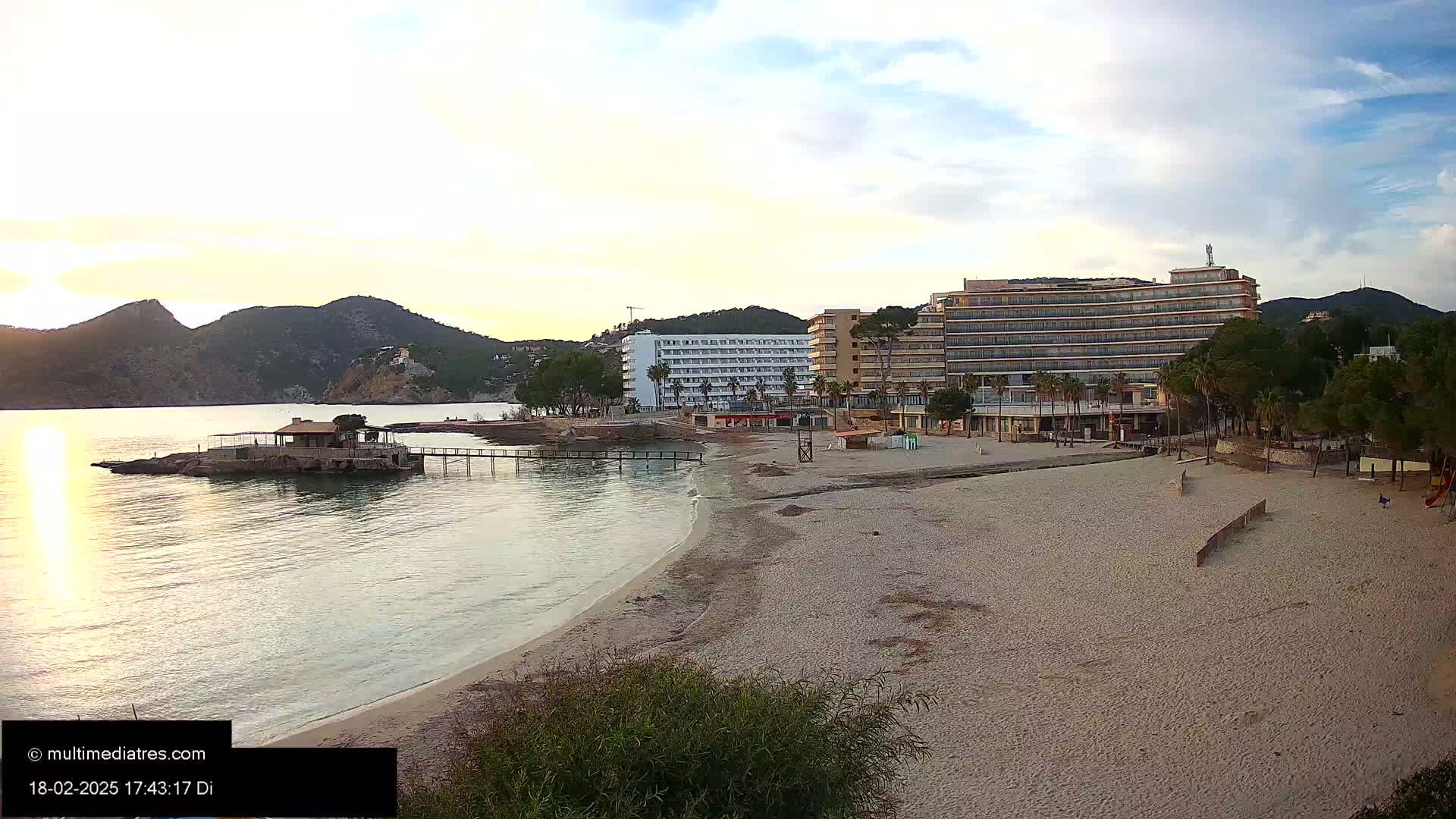 A sandy beach with calm water, hotels, and palm trees under a partly cloudy sunset sky.