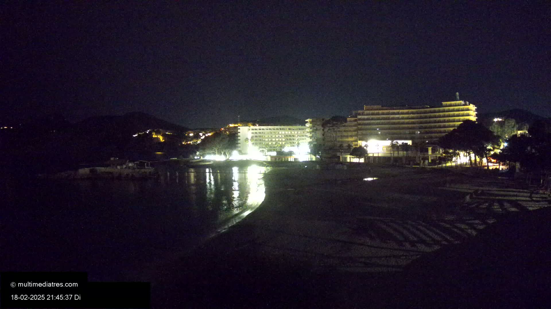 At night, illuminated hotels are situated near a dark beach and calm body of water, with a hill visible in the background.