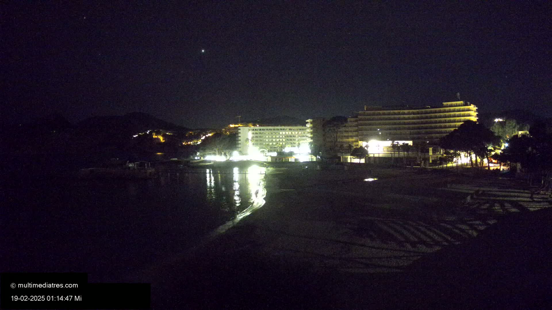 A dark, night-time view of a beach with two illuminated hotels and a calm body of water reflecting their lights.