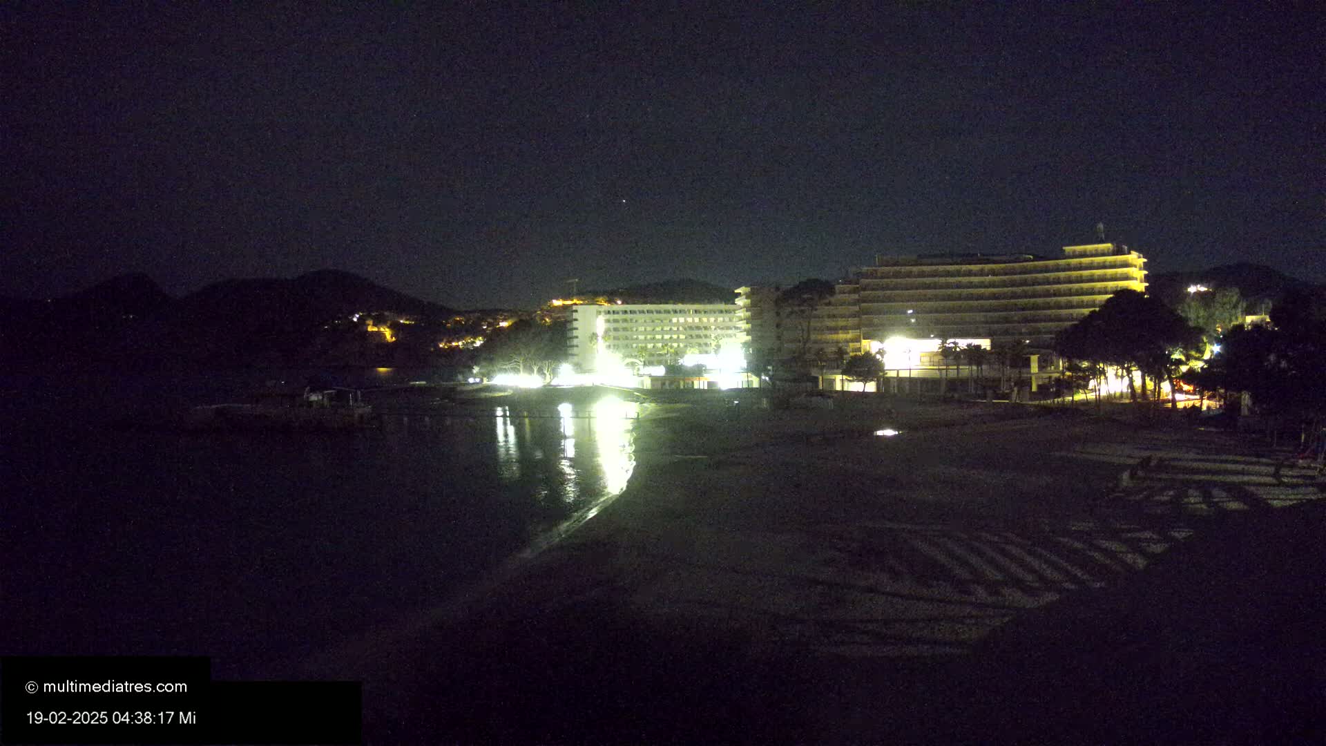 At night, two illuminated hotels are situated on a dark beach next to a calm body of water and a hill.