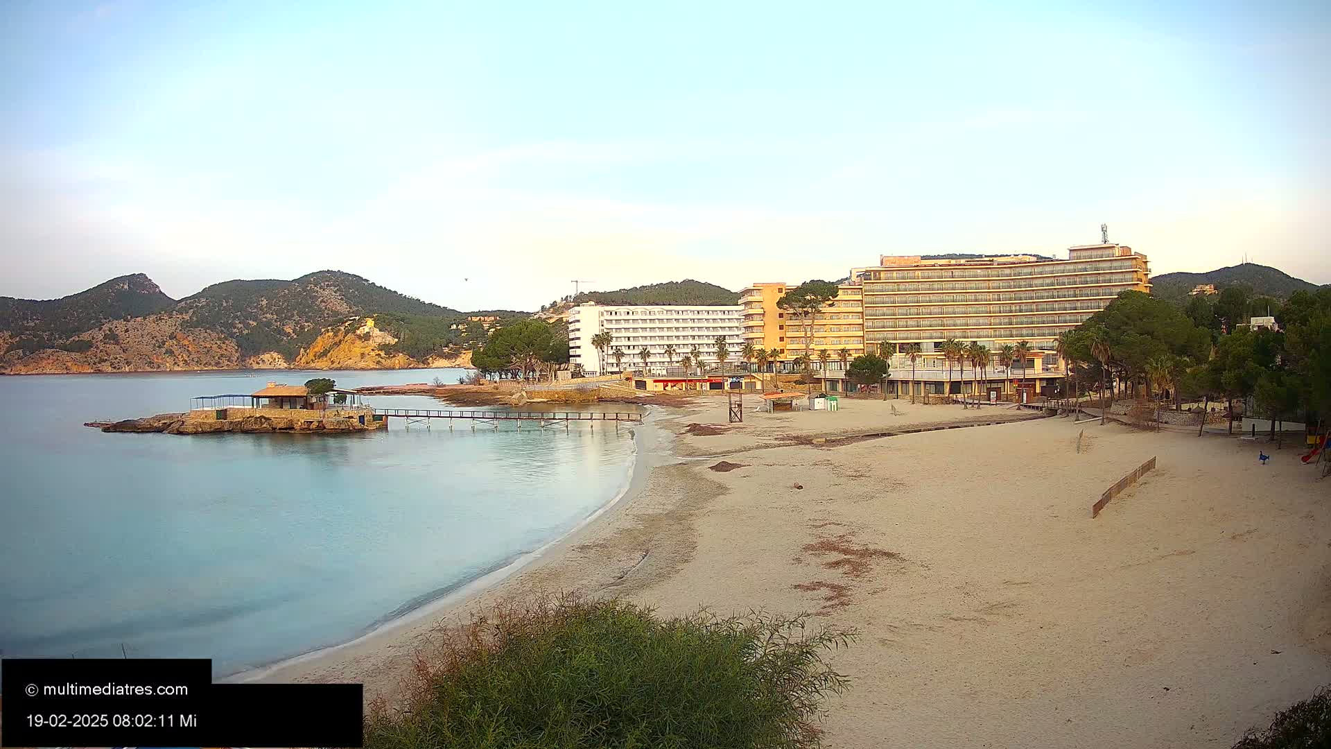 A sandy beach with calm, clear water stretches out before two large hotels and a small, stone building on a small island, all set against a backdrop of hills under a clear, light sky.