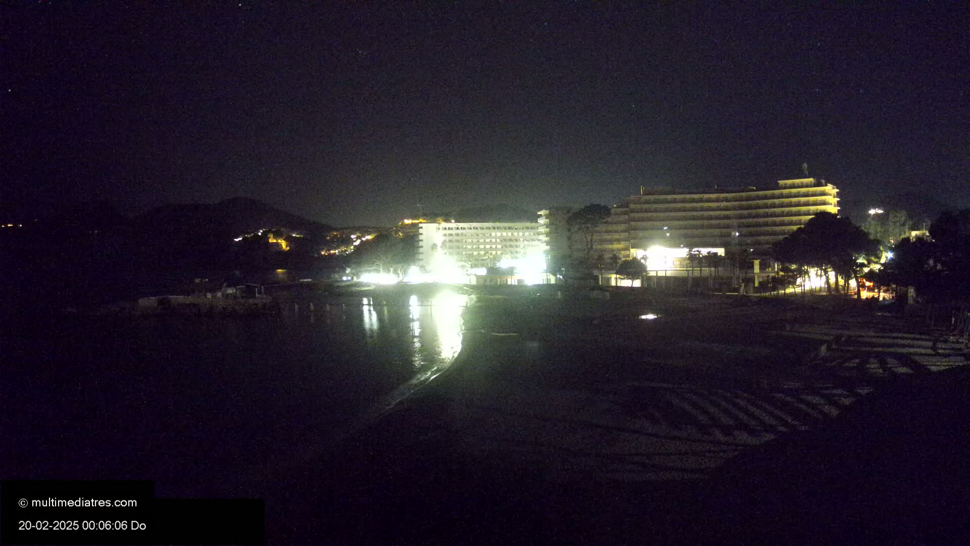 Two brightly lit hotels are visible on a dark beach at night under a clear, starry sky.