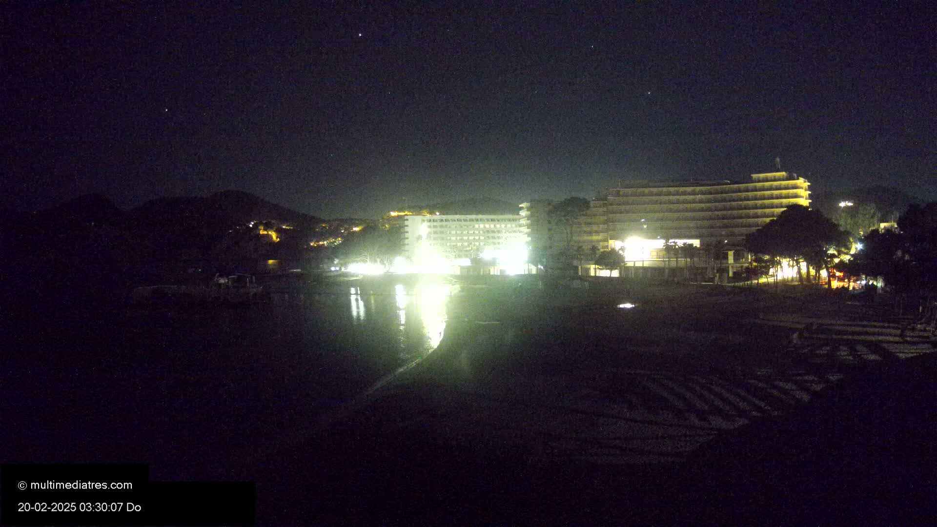 At night, two brightly lit hotels are situated on a dark beach near a hill under a clear, starry sky.