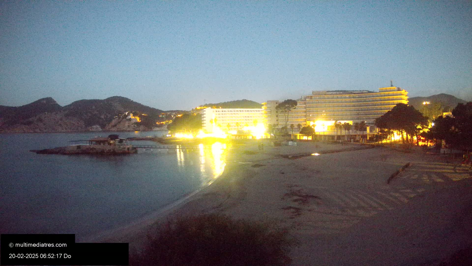 A sandy beach at twilight, with illuminated hotels and a small island visible across calm water, under a clear, dark sky.