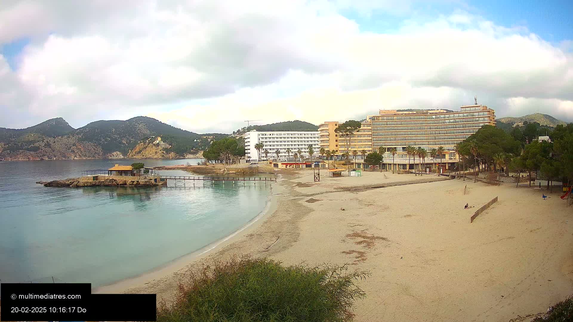 A sandy beach with calm, clear water curves toward two hotels situated beside a small cove backed by a mountainous region under a partly cloudy sky.