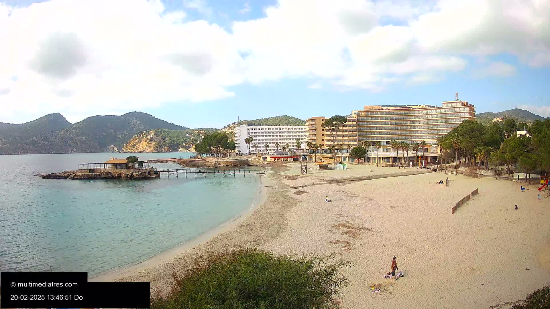 A sandy beach with a few people, a small island with a structure, and large hotels behind it, all under a partly cloudy sky.
