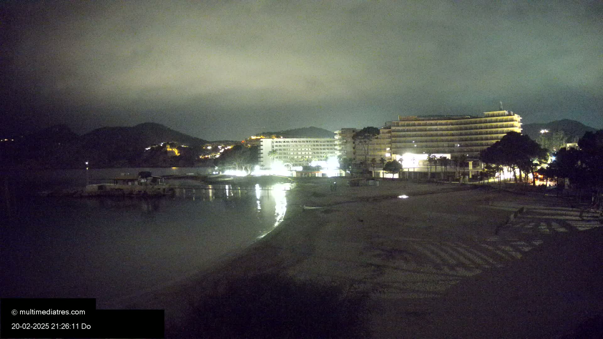 A dark, nighttime beach scene shows two illuminated hotels near a calm body of water under a cloudy sky.