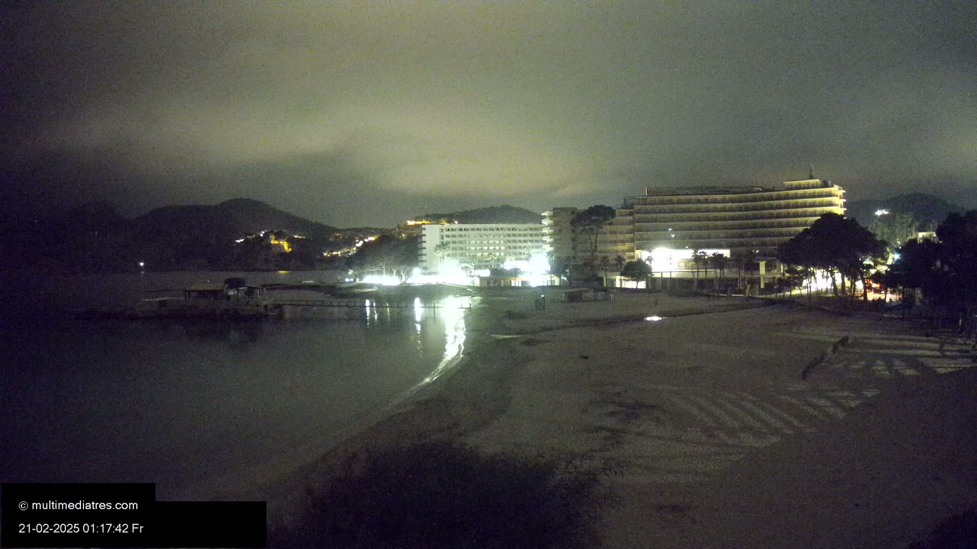 At night, under a cloudy sky, two illuminated hotels are situated near a dark beach and calm body of water with a mountain in the background.