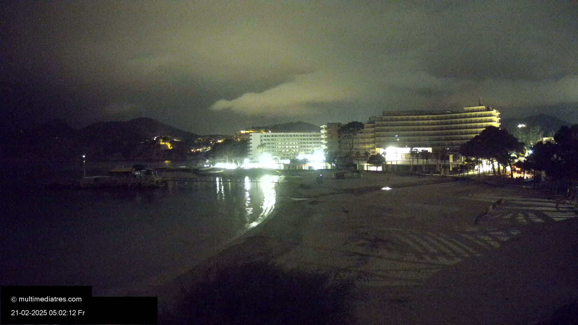 At night, under a cloudy sky, illuminated hotels are situated on a beach next to a calm body of water with a mountain in the background.
