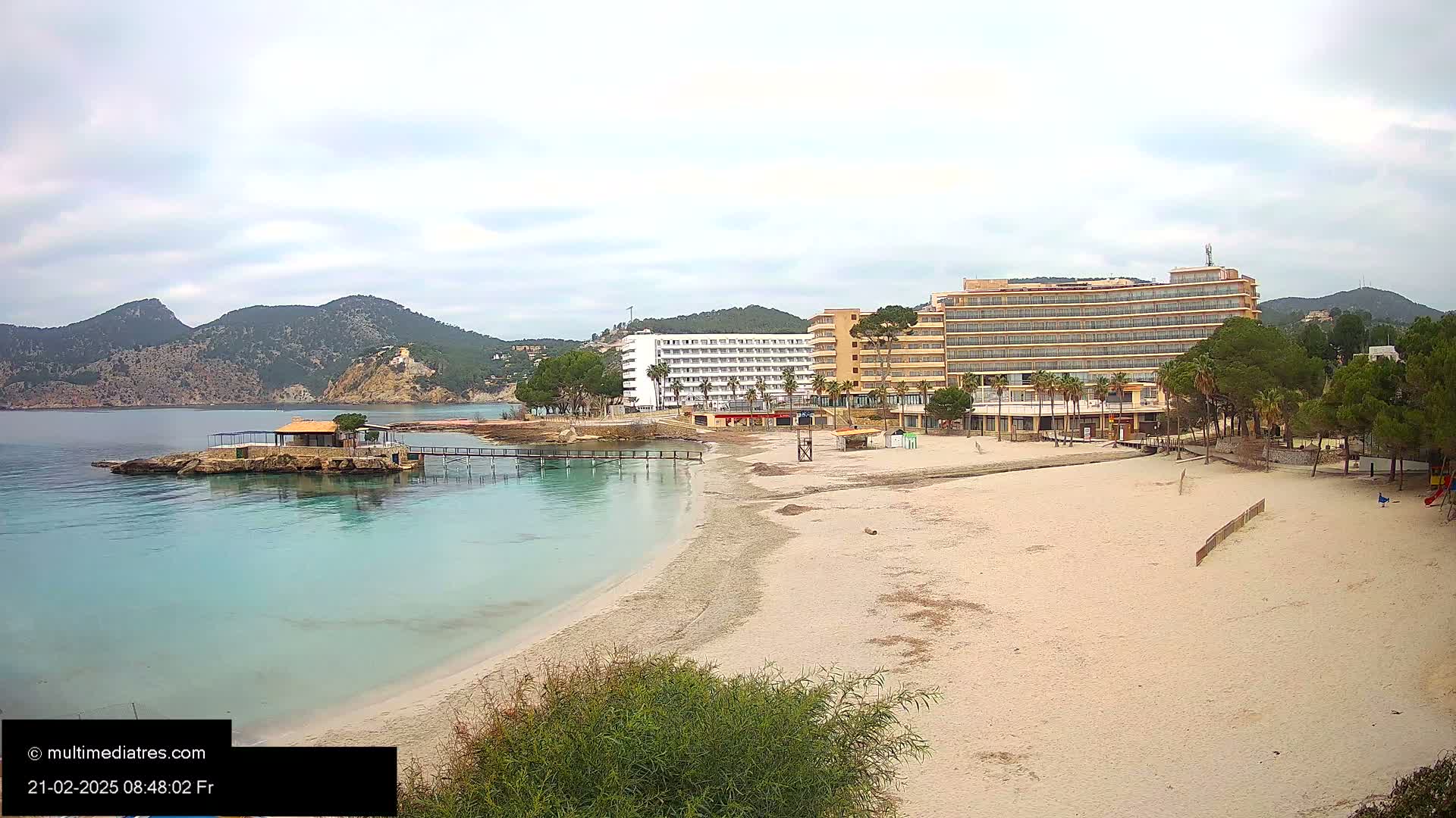 A sandy beach with clear, turquoise water and two hotels sits at the base of a mountain range under an overcast sky.