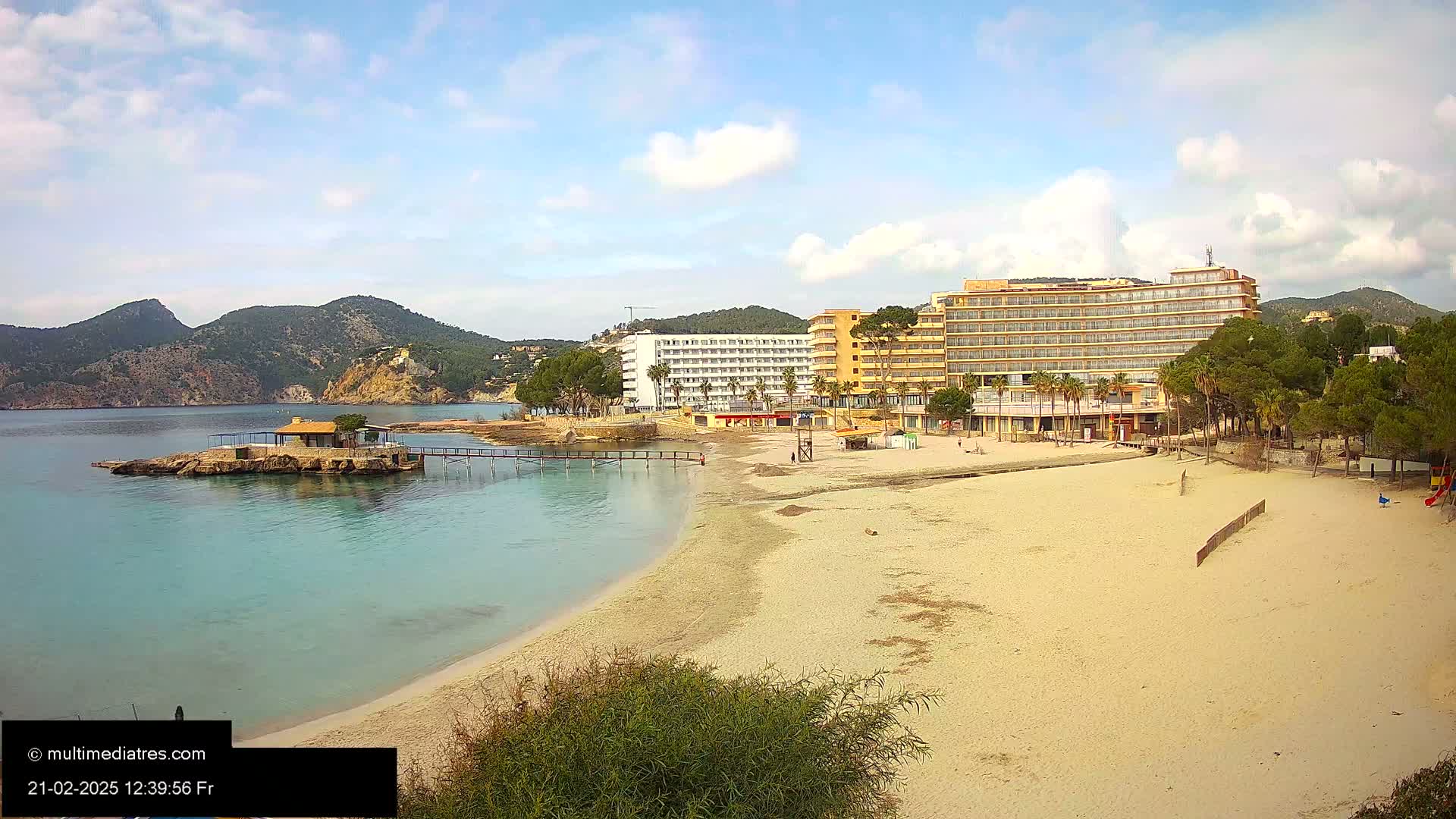 A sandy beach with calm, turquoise water stretches toward hotels and green hills under a partly cloudy sky.