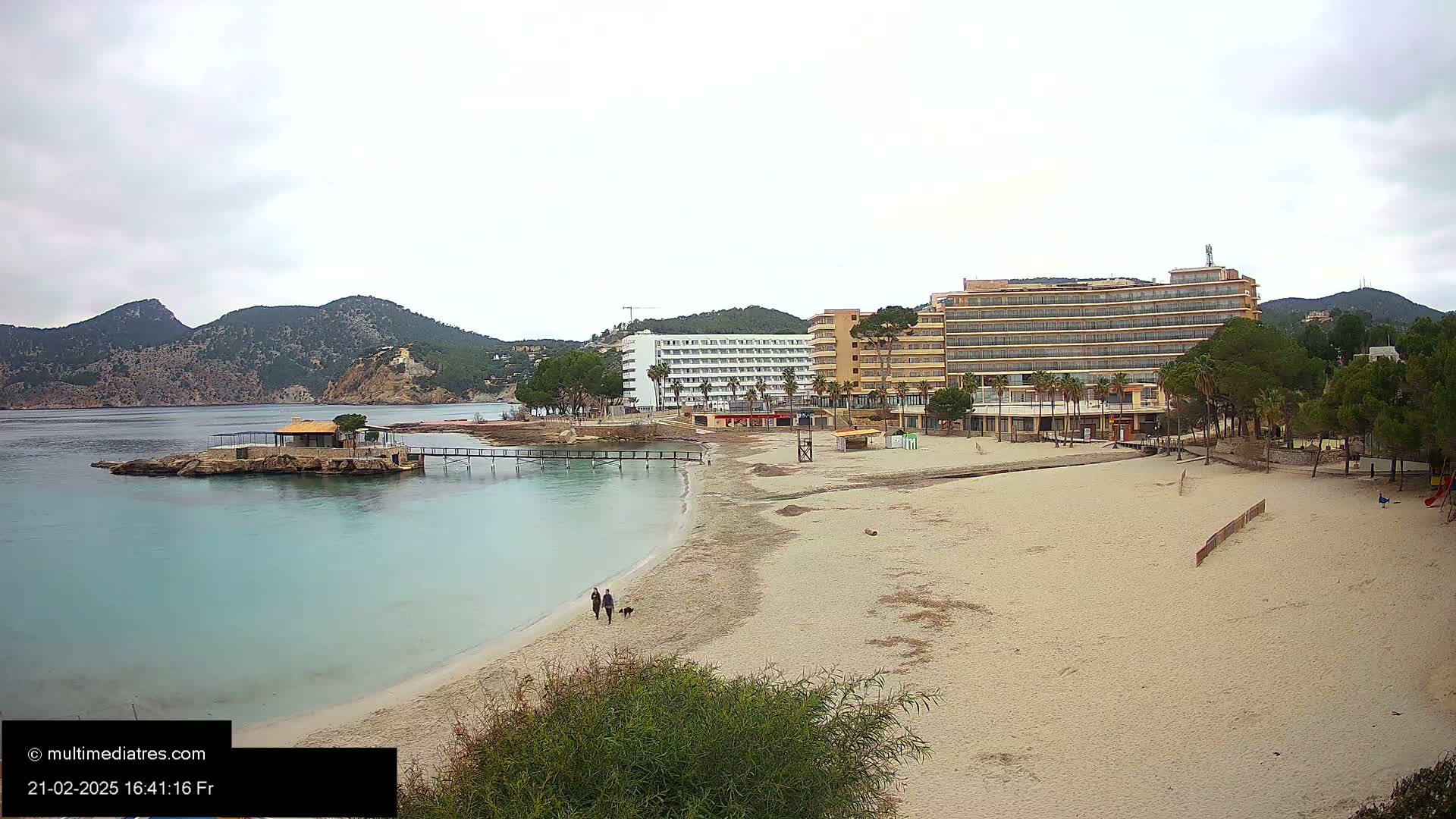 A sandy beach with two hotels and a small island in calm, overcast water, backed by mountains.