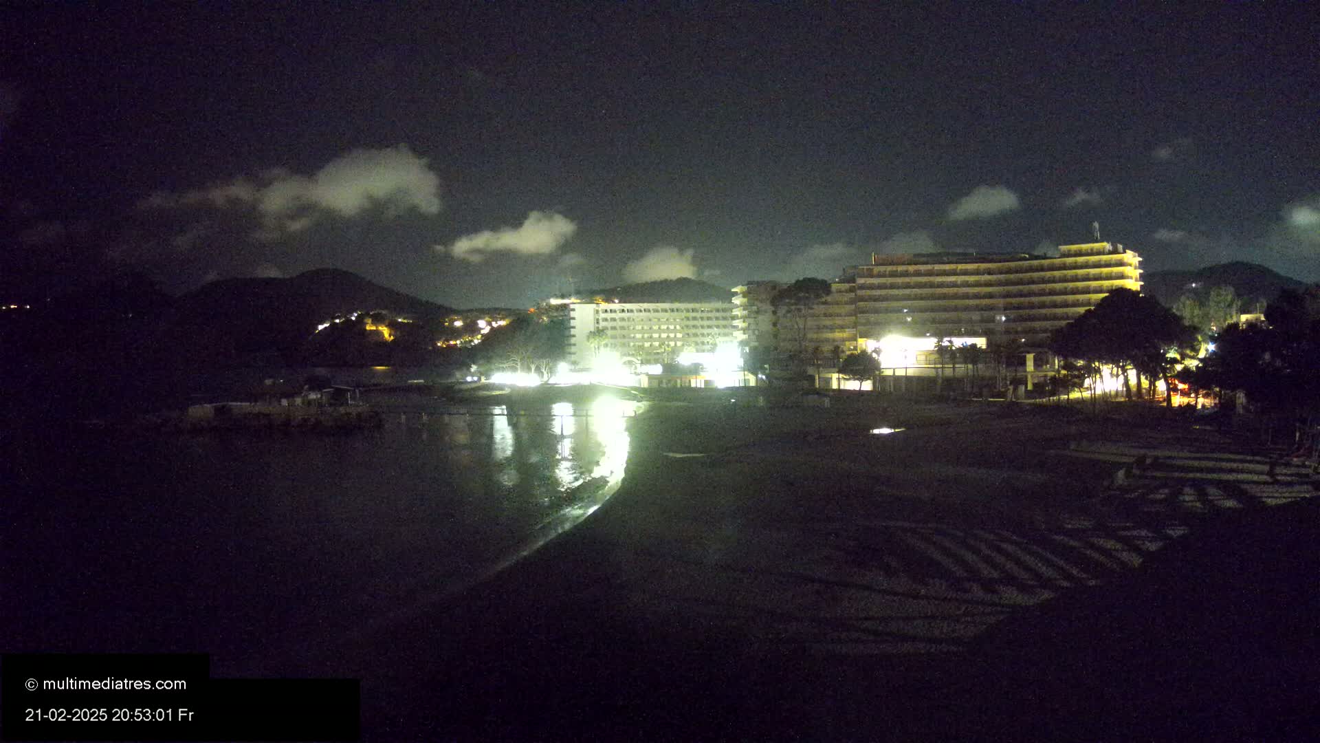 At night, under a partly cloudy sky, illuminated hotels and buildings are situated near a dark beach and calm body of water, backed by a hill.