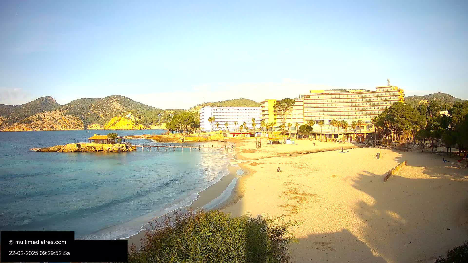A sandy beach with calm, clear turquoise water laps at its shore, backed by hotels and low-lying hills under a clear blue sky.