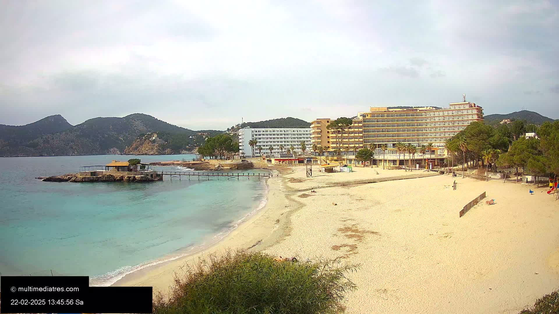 A sandy beach with calm, turquoise water stretches out in front of several hotels and a small island with a structure on it, under an overcast sky.
