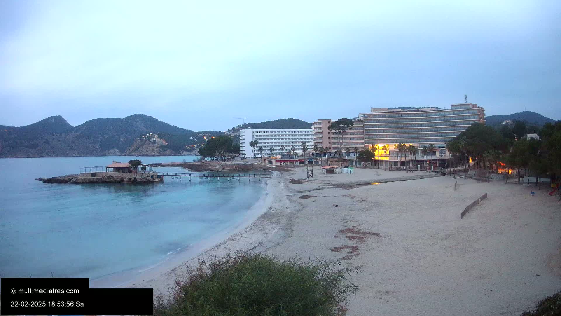 A sandy beach with calm, blue-green water, two hotels, and a small island with a building are visible under a twilight sky.