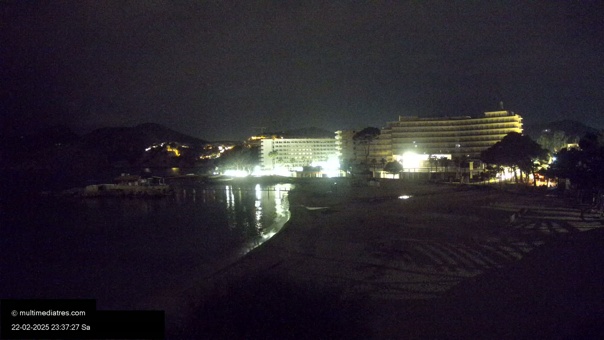 At night, two brightly lit hotels stand on a dark beach near a calm body of water, with a hill visible in the background.