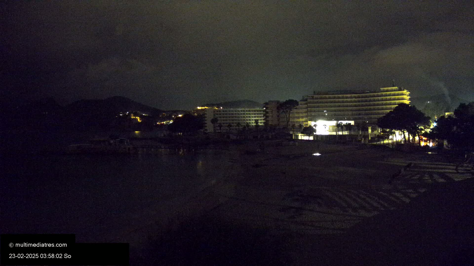 A dark, nighttime beach scene with dimly lit hotels and a hill in the background under a cloudy sky.