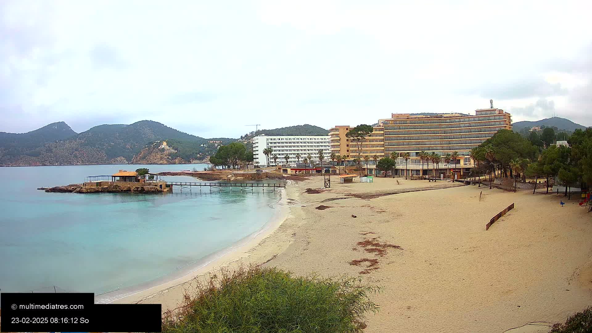 A sandy beach with calm turquoise water and two hotels is seen under an overcast sky, backed by low, green-brown mountains.