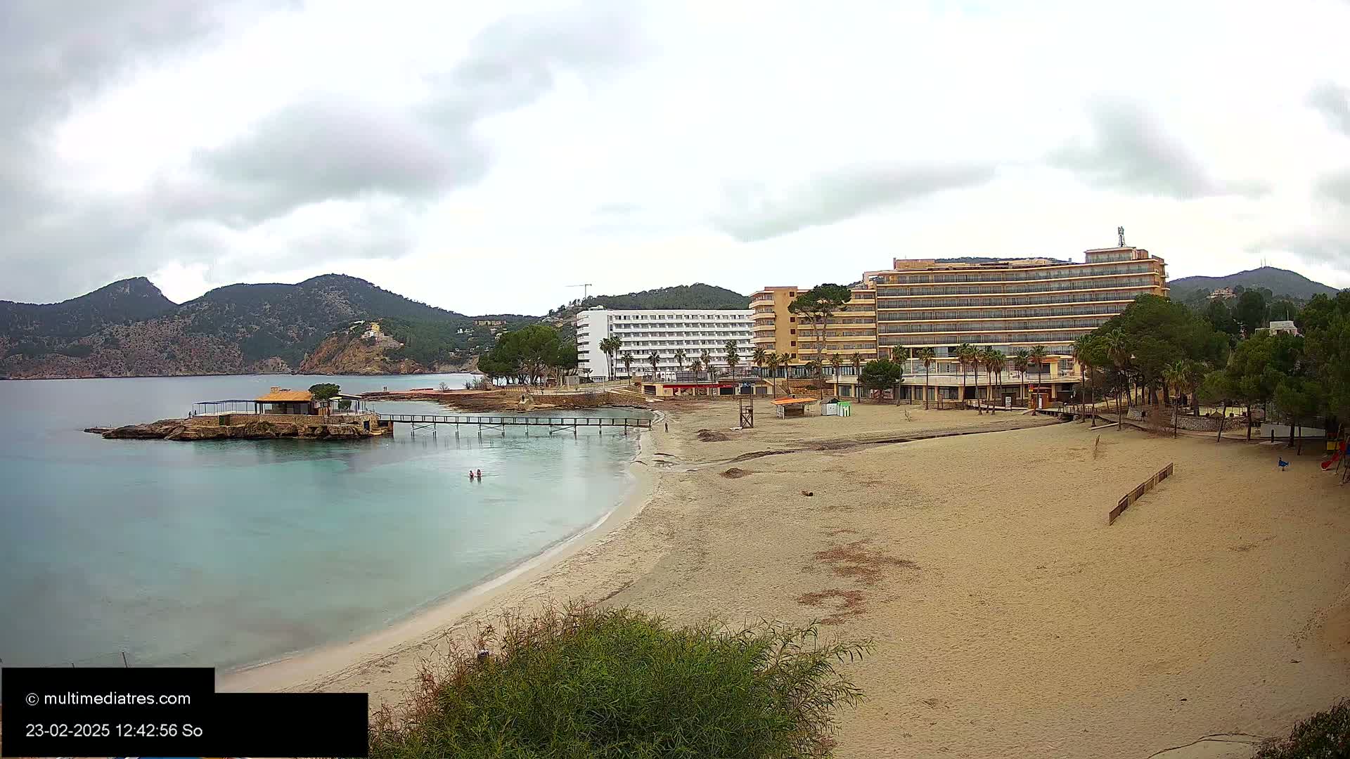 A sandy beach with two hotels and a small island with a building is visible under an overcast sky.
