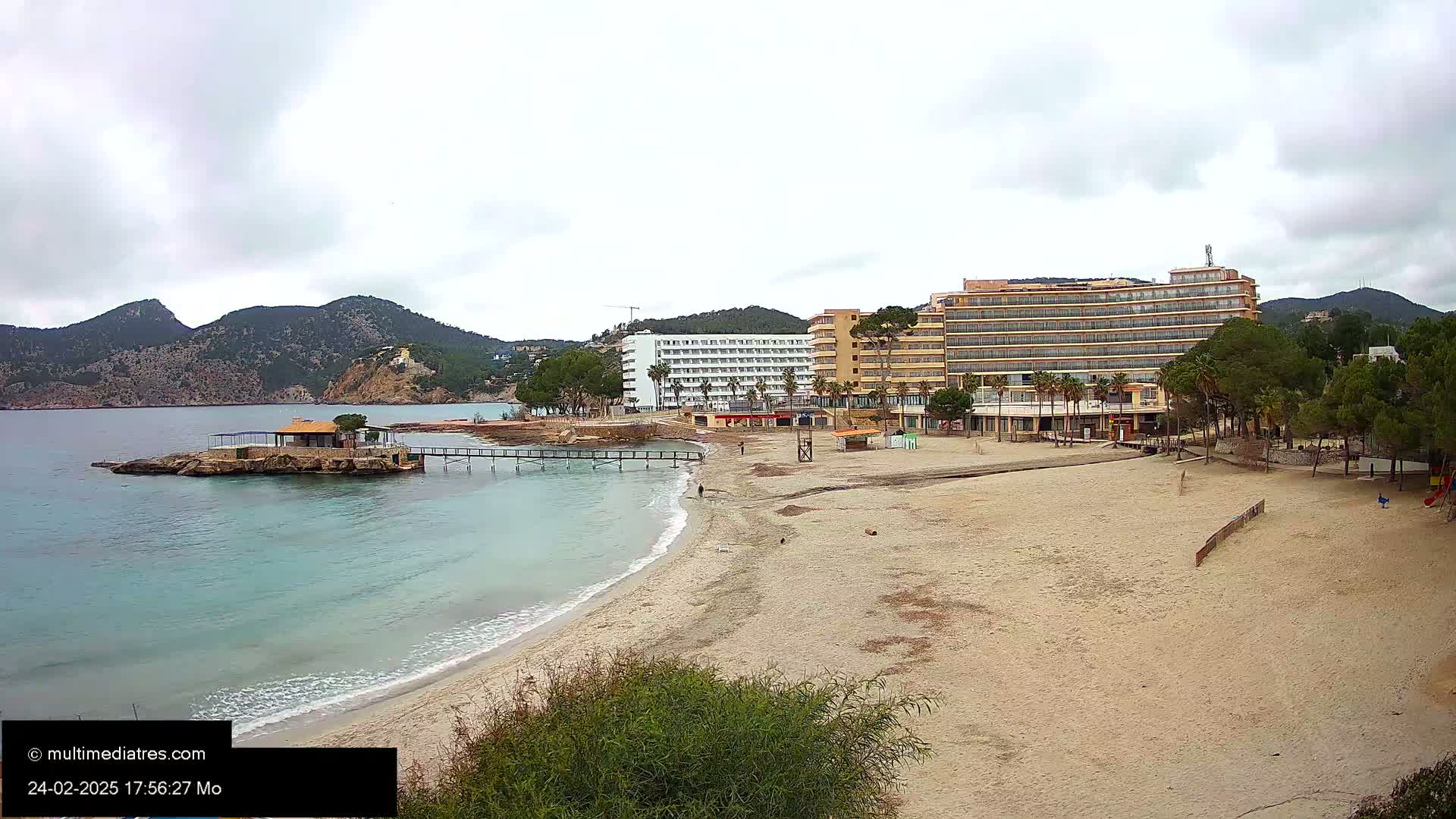 A sandy beach with calm, light-blue water stretches toward two hotels and a small island with a building, all situated against a backdrop of mountains under an overcast sky.