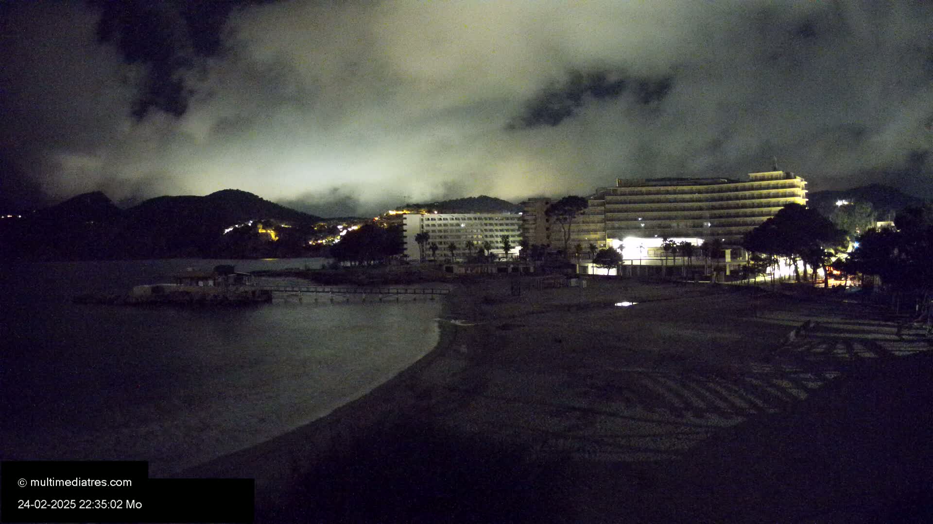 A dark, cloudy night scene shows a beach with several lit hotels and a small structure in the water, backed by a mountain range.