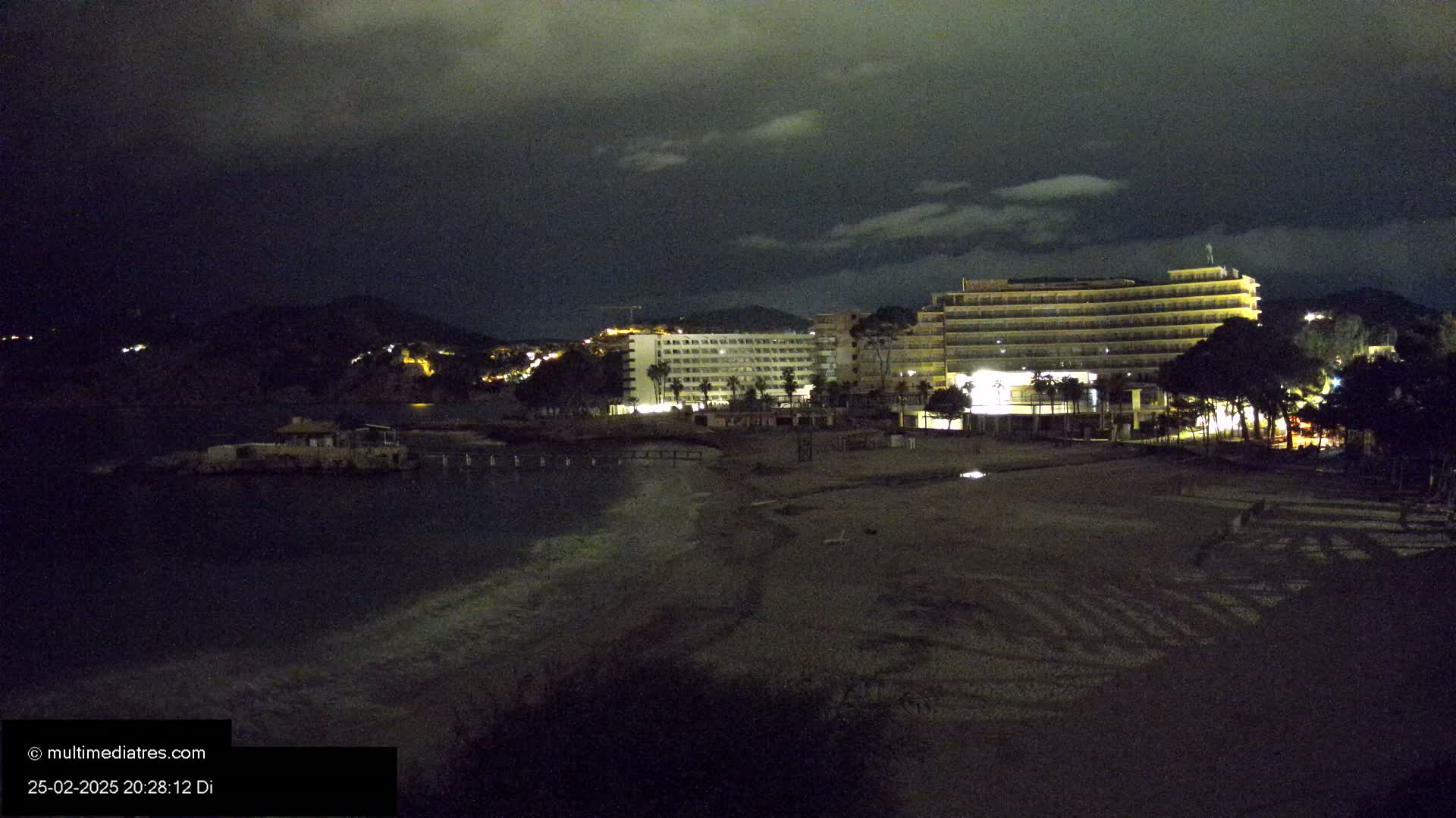 At night, under a dark, cloudy sky, a beach with dimly lit hotels and a few trees sits beside a dark body of water.
