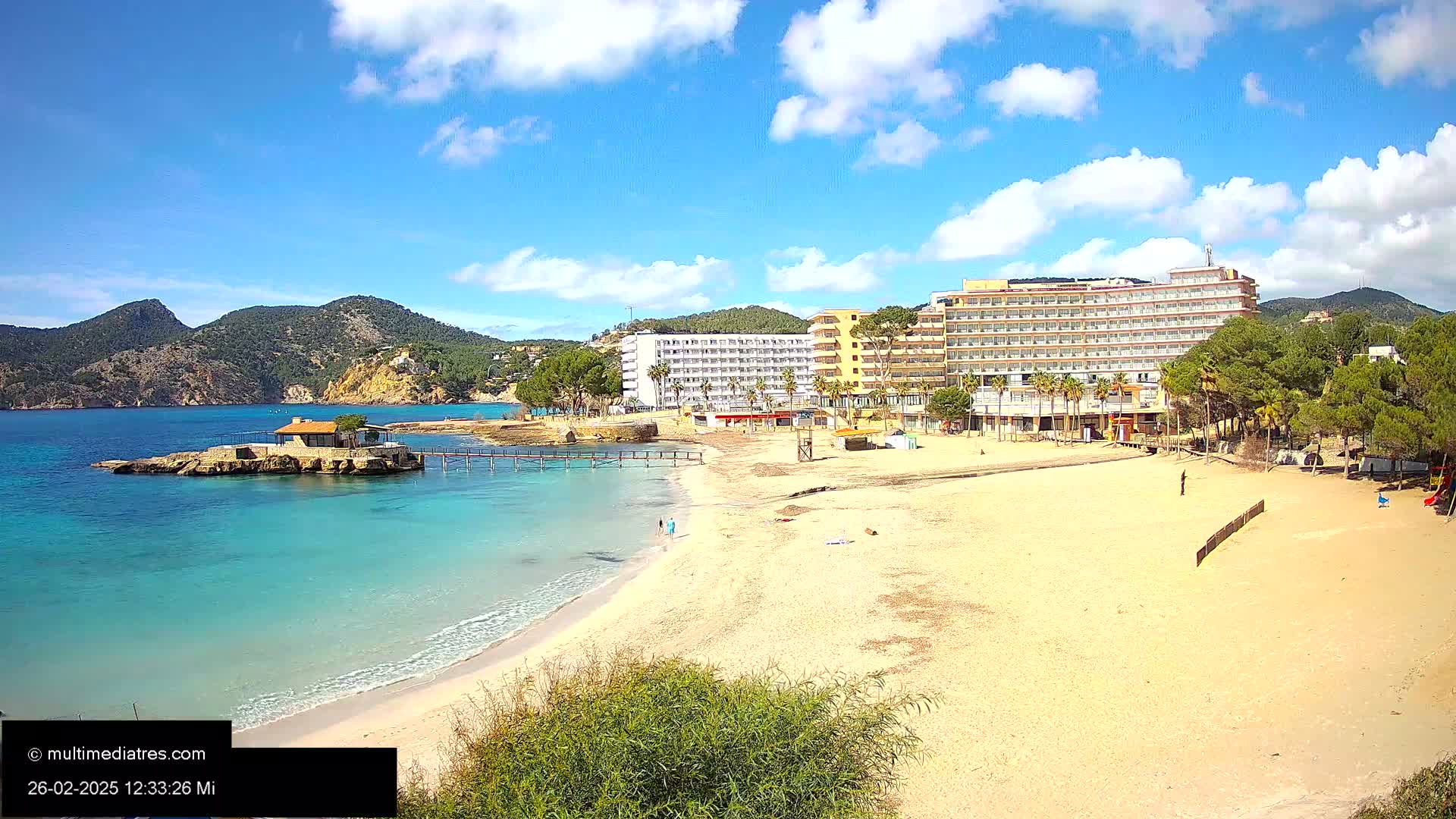 A sandy beach with turquoise water, a small island with a building, and large hotels behind it, all under a mostly sunny sky with some clouds.
