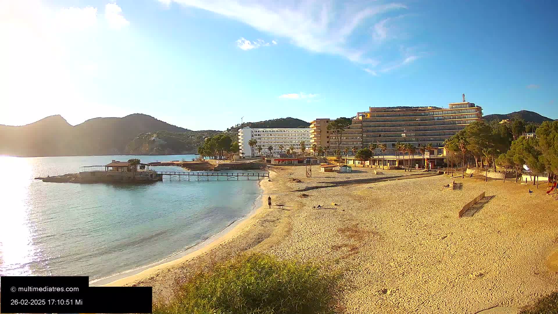 A sandy beach with a few people on it stretches out in front of several hotels and a line of trees, all under a bright, sunny sky.