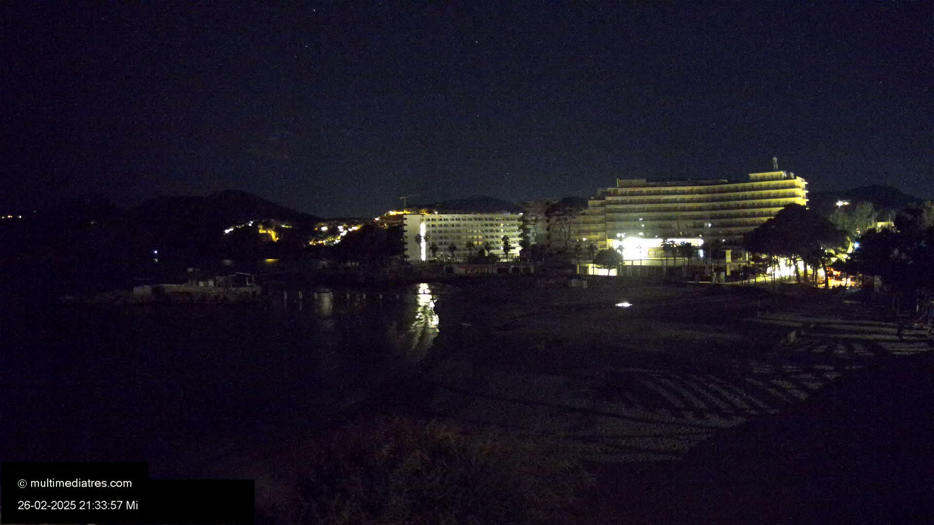 A nighttime view of a beach with illuminated hotels and a hill in the background under a clear, starry sky.