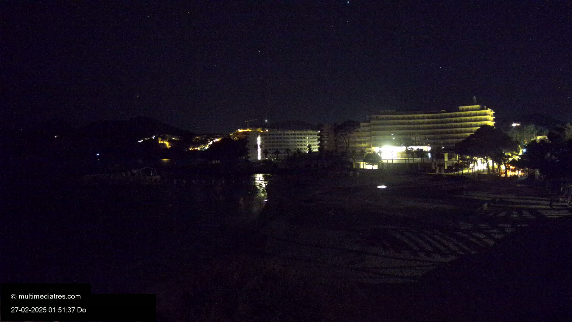 At night, under a clear, starry sky, illuminated hotels and buildings are situated near a dark beach.