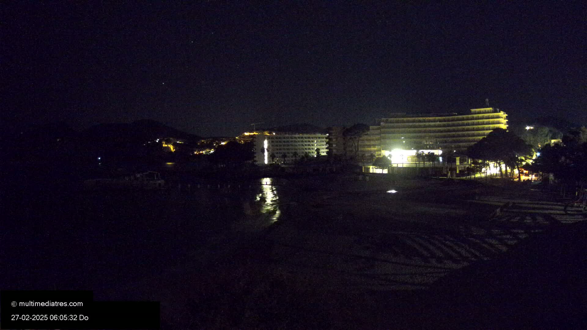 A nighttime view of a beach with illuminated hotels and a hill in the background under a clear, starry sky.
