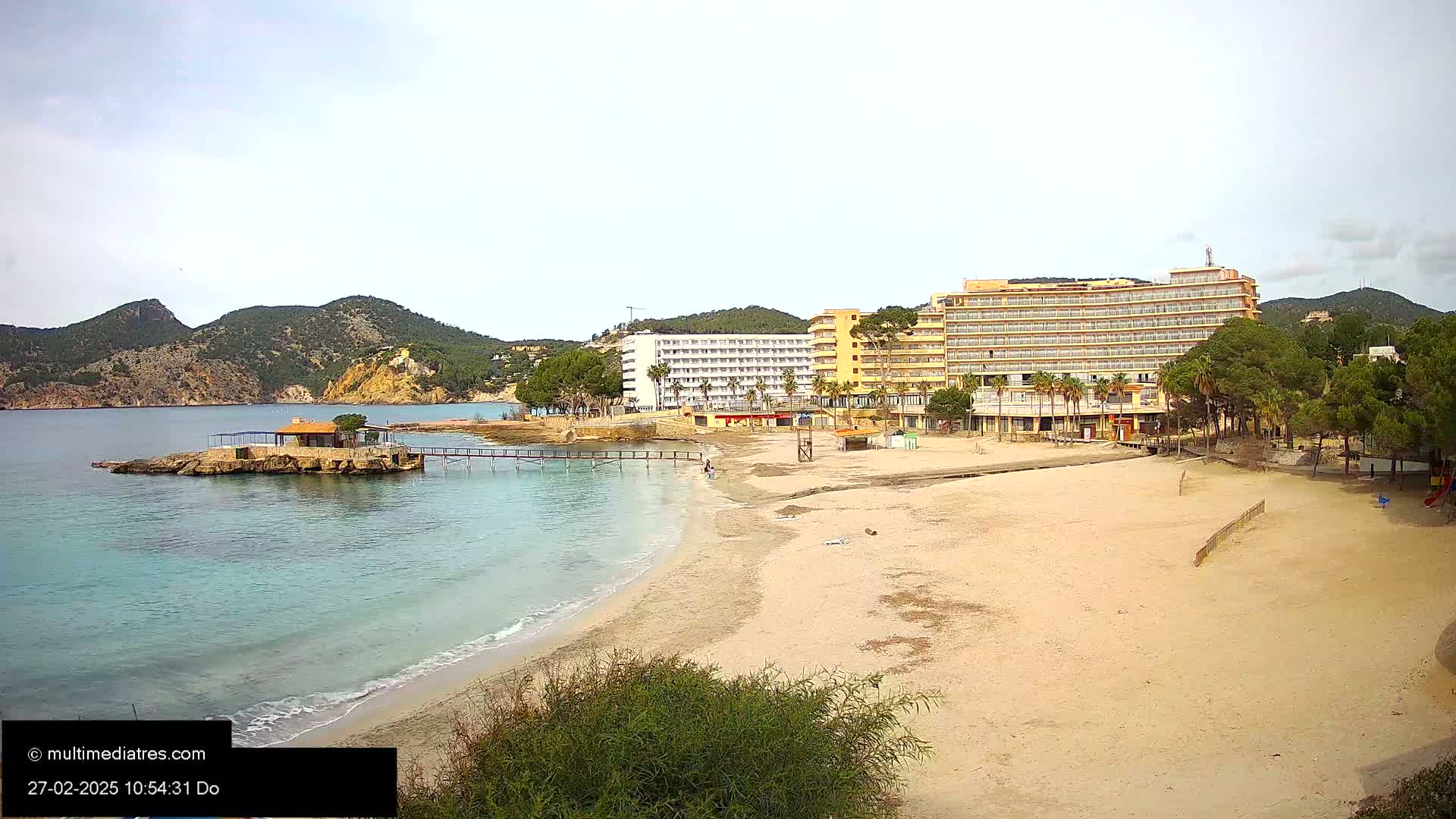 A sandy beach with calm, clear water stretches towards two large hotels and a small island with a structure, all set against a backdrop of green hills under an overcast sky.