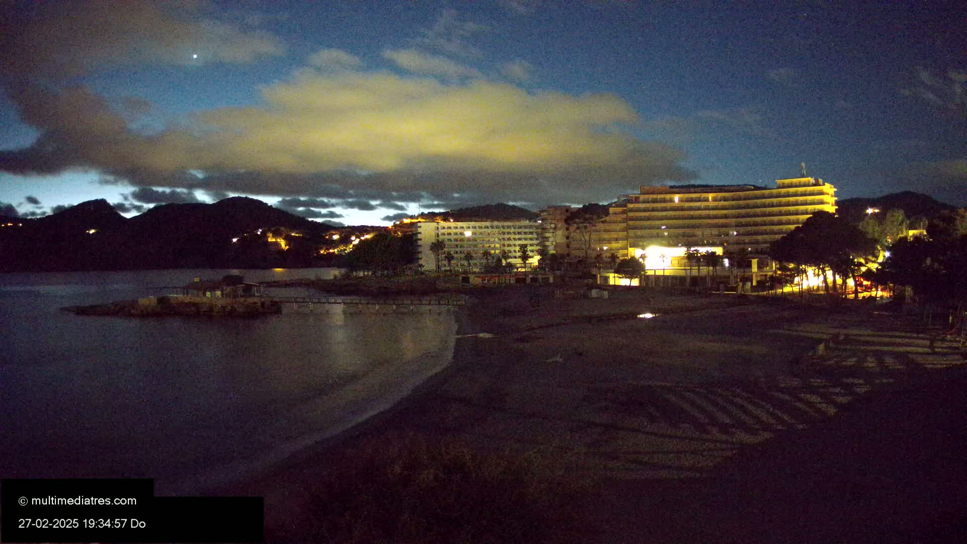 A dark, nighttime beach scene shows a mostly-dark shoreline, a calm body of water, and illuminated hotels and buildings behind it, under a cloudy sky.