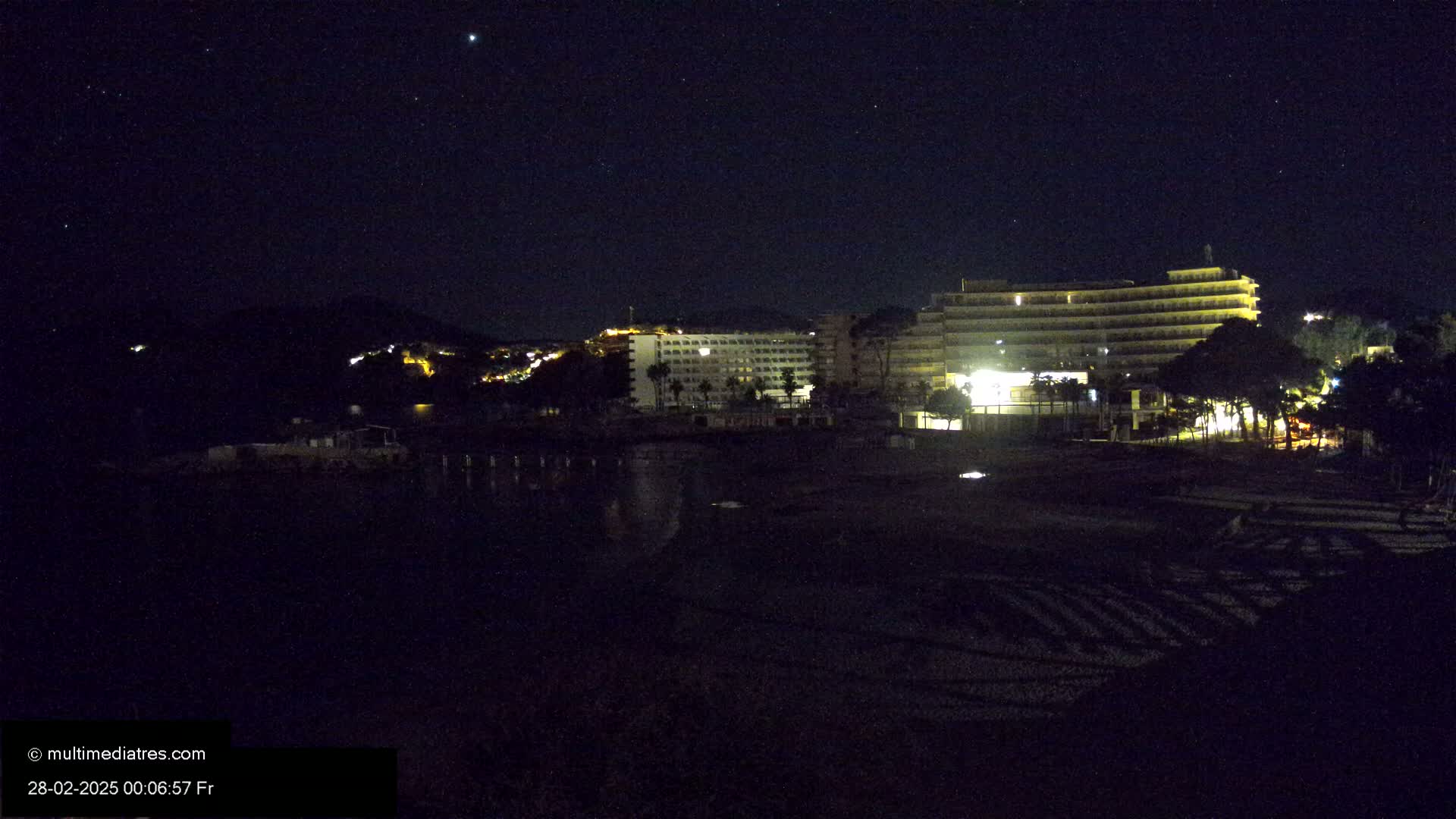 A nighttime view of a beach with illuminated hotels and a hill in the background under a clear, starry sky.