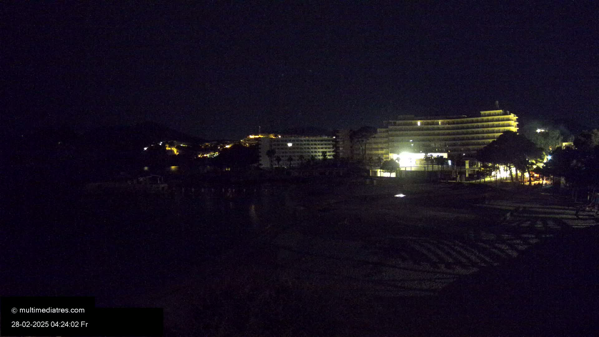 A dark, nighttime beach scene shows several illuminated hotels and buildings across a body of water, under a clear, starlit sky.