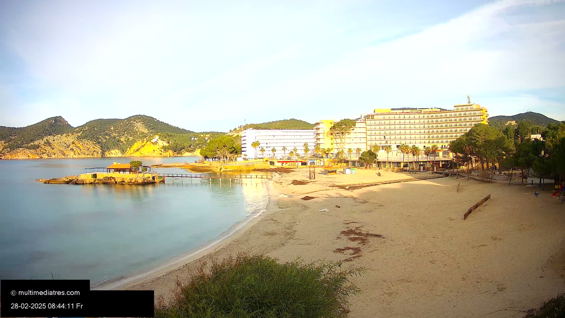 A sandy beach with calm, clear water stretches out before a row of hotels nestled against hills under a clear, bright sky.