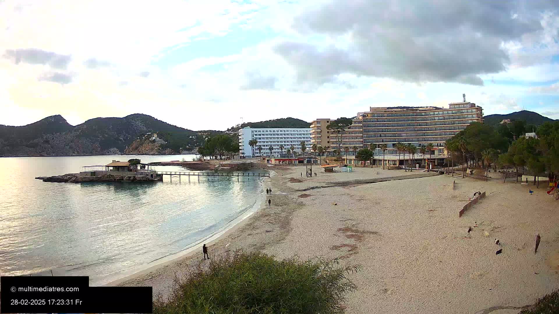 A sandy beach with several people on it is situated near a calm body of water, backed by hotels and mountains under a cloudy sky.