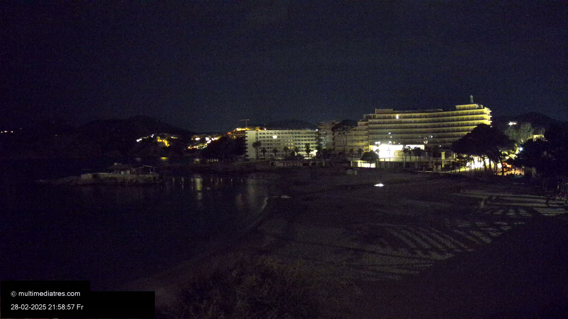 A nighttime view of a beach with illuminated hotels and a calm body of water in the foreground, under a dark sky.