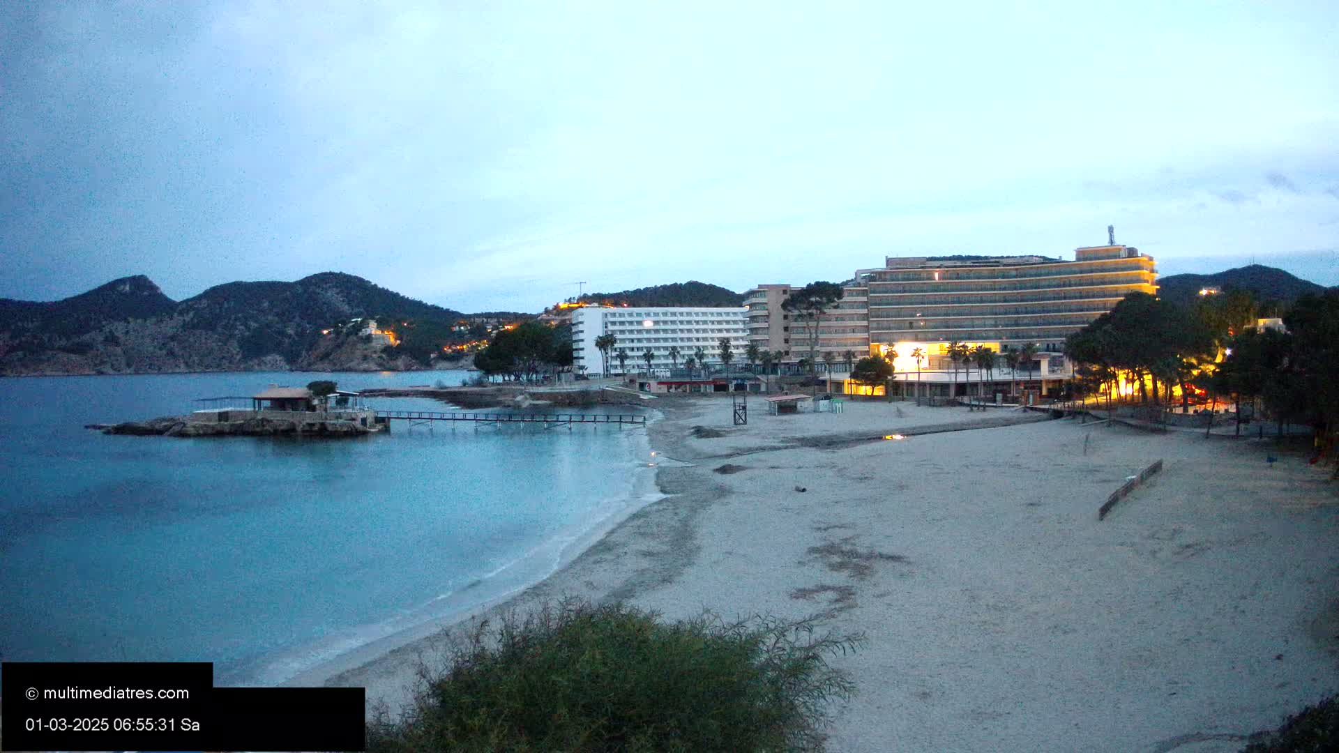 A sandy beach at dusk, with calm, blue water, hotels, and trees, under a twilight sky.