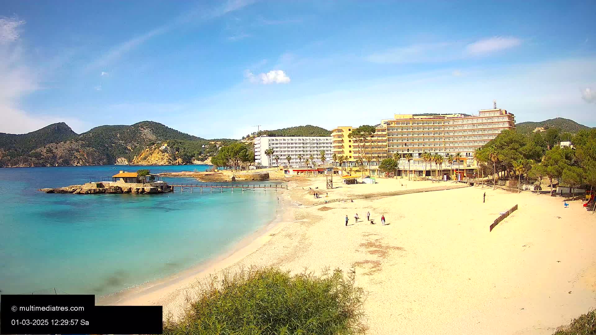 A sandy beach with turquoise water, a small building on a rock, a large hotel, and a few people are visible under a sunny sky.