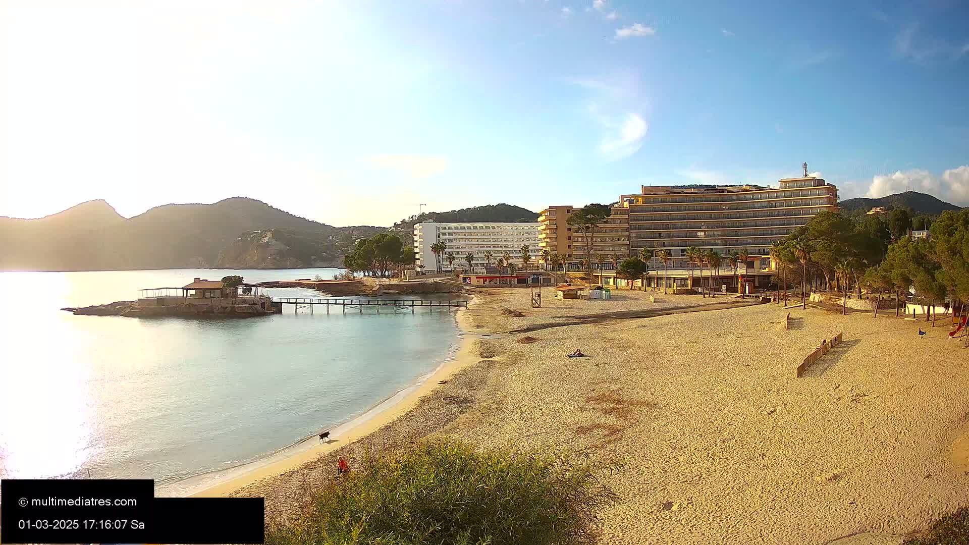 A sandy beach with calm, clear water laps at its edge, backed by hotels, palm trees, and hills under a mostly sunny sky.